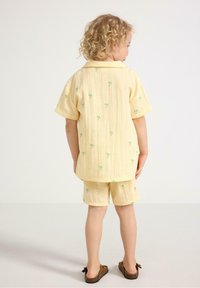 Gul t-shirt med kort ärm och shorts-set med små gröna palmer broderade, tillverkat av ett texturerat, lätt tyg. Bruna slip-on skor.