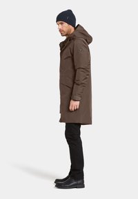 Ruskea vedenpitävä parkatakki hupulla, tumma neulottu pipo, mustat slim-fit farkut ja mustat saappaat. Sileä pinta ja minimalistinen muotoilu.