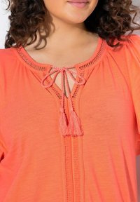 Korálový top s krátkým rukávem s detaily zavazování a třásněmi, složitým vyšíváním výstřihu a volným střihem. Měkká, lehká látka.