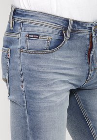 Gai zila denima bikses ar klasisku piecu kabatu dizainu, taisnu kāju, kontrastējošo vīlēšanu un zīmola uzlīmi uz jostas.