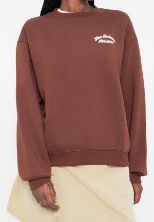 Persoon die een bruine New Balance Athletics trui en een lichtbeige rok draagt, staand tegen een effen achtergrond.