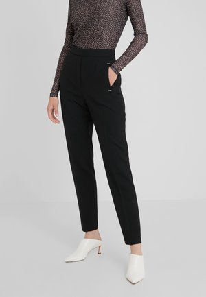 Persoon die zwarte, hooggetailleerde taps toelopende broek, een patroonrijke top met lange mouwen en witte, met hakken uitgevoerde muilezels draagt, staand met één hand in de zak.