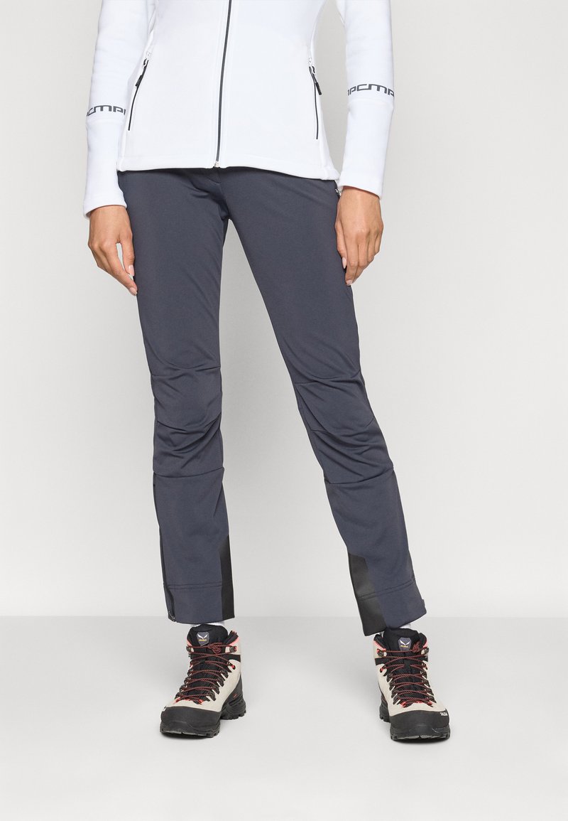 Persona che indossa pantaloni outdoor grigio scuro, scarpe da trekking con lacci rossi e una giacca bianca con zip e tasche con zip.