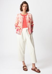 Floral-patterned cardigan in pink and orange, layered over a coral top, paired with ivory trousers and brown sandals. Lightweight fabric, relaxed fit.