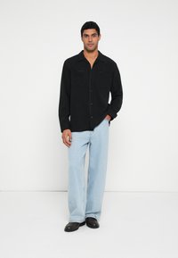 Musta napillinen paita, jossa on kaksi rintataskua, yhdistetty vaaleansiniset leveälahkeiset farkut ja mustat kengät, yksinkertaisella valkoisella taustalla.