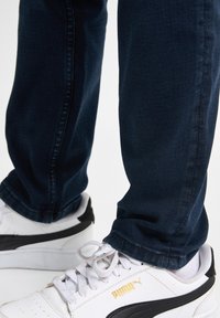 Zapatillas blancas con acentos negros y branding dorado, combinadas con jeans de mezclilla azul oscuro. Los jeans tienen un corte ajustado y detalles de costura visibles.