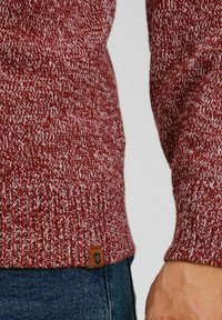 Pull en tricot rouge et blanc à motifs, avec des poignets côtelés, agrémenté d'une petite étiquette en cuir marron à l'ourlet, porté sur un jean bleu.