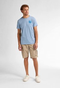 Vaaleansininen puuvillainen t-paita pyöreällä pääntiellä ja rintakehän grafiikkaprintillä, yhdistettynä beigenvärisiin cargoshortseihin ja valkoisiin lenkkareihin.