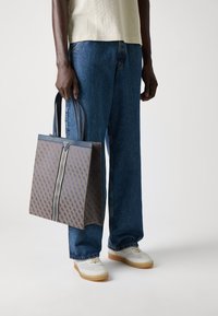 Sac fourre-tout en toile marron avec un motif géométrique, doté de poignées en cuir bleu et d'une bande centrale. Le modèle tient le sac tout en portant un jean.