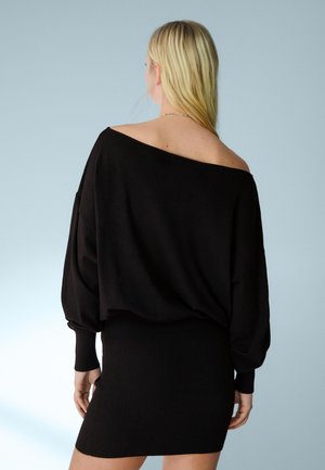 Femeie blondă văzută din spate, purtând un top negru cu mâneci lungi, desprins de pe umeri, și o fustă neagră cu dungi, pe un fundal simplu, deschis la culoare.