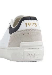 Witte sneaker met grijs suède en zwarte leren accenten, gouden "1973" detail op de hiel, en een gestructureerde rubberen zool met het merksymbool.