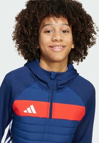 Mornarska sportska dukserica od teksturirane tkanine s crvenom prugom preko prsnog dijela, s patentnim zatvaračem i bijelim logotipom s lijeve strane.