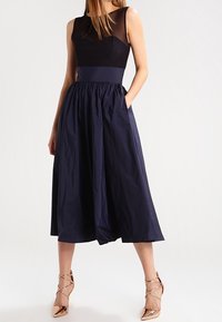 Schwarz und marineblaues Kleid mit tailliertem Oberteil, fließendem knielangem Rock und Taschen. Glattes Material mit leichtem Glanz. Metallische Sandalen mit Riemchen.