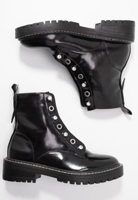 Černé lakované kotníkové boty s kulatými špičkami, ozdobené hřebíky a s tlustou gumovou podrážkou. Obsahují poutko na zadní straně.