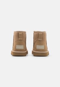 Botas de tobillo de ante marrón con un suave forro interior, suela de goma y una lengüeta en la parte trasera. Etiquetadas con un logo UGG gris claro en el talón.