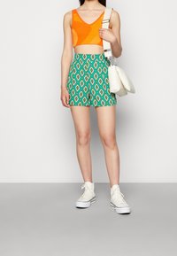 Cropped top a fantasia arancione e gialla, pantaloni verde con un motivo a diamante, borsa bianca e sneakers alte bianche.