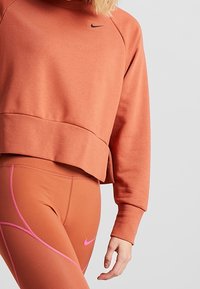 Sudadera recortada de Nike en un naranja apagado, con un logo negro, combinada con leggings a juego que tienen detalles en rosa y un patrón texturizado.