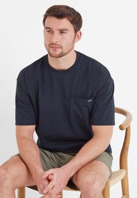 Navy blue short-sleeve t-shirt made of cotton, featuring a chest pocket and a relaxed fit. Model sits on a wooden chair.