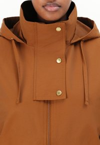 Veste marron imperméable avec un col haut, dotée d'une fermeture éclair sur le devant et de quatre boutons-pression dorés. Texture lisse avec cordons de serrage ajustables.