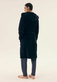 Czarny, pluszowy szlafrok z kapturem, z paskiem w talii. Noszony na granatowych spodniach piżamy, z odsłoniętymi stopami. Minimalistyczne tło.