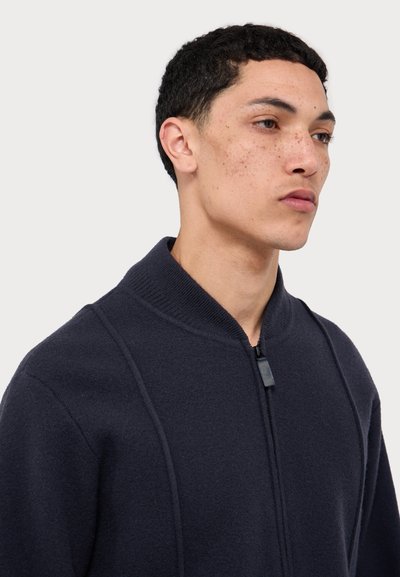 Tamsiai mėlyna zip-up megztinis, pagamintas iš minkštos medžiagos. Jame yra aukšta apykaklė, subtilūs siuvimo akcentai ir lygus zip-pull.
