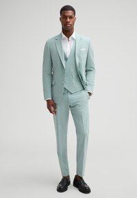 Light green three-piece suit made of textured fabric, featuring a fitted blazer, coordinating waistcoat, and tailored trousers. Black loafers.