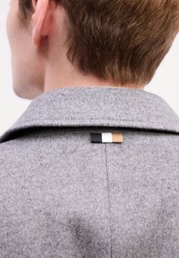 Cappotto di lana grigio con colletto a punta. Sul retro presenta un piccolo cartellino in nero, bianco e beige, attaccato alla cucitura del collo. Texture morbida visibile.