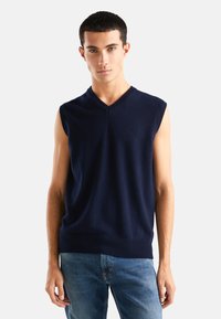 Granatowy bezrękawnik z dekoltem w serek, wykonany z miękkiej dzianiny, o dopasowanym kroju i prążkowanym wykończeniu. Nosi się go na jasnoniebieskich dżinsach.