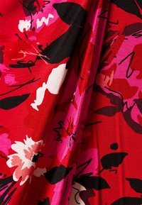 Tissu rouge avec imprimé floral abstrait en noir, blanc, rose et rouge, montrant des plis et des froissements dans le matériau.