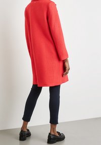 Red wool coat with a relaxed fit, long sleeves, and a back vent. Paired with navy leggings and black platform shoes, viewed from the back.
