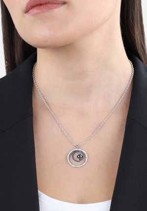 Zilveren ketting met geconcentreerde cirkelvormige hangers, een centrale kraal en kleine heldere stenen. Draagt tegen een donkere blazer.