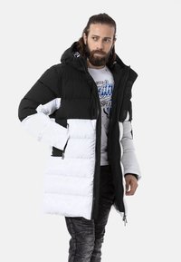 Schwarze und weiße gepolsterte Jacke mit Kapuze, ausgestattet mit Reißverschlusstaschen und einer strukturierten Oberfläche. Der schwarze obere Teil kontrastiert mit dem weißen unteren Teil.