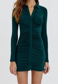 Robe verte à manches longues en tissu doux avec un devant boutonné, un design ajusté et des accents froncés sur les côtés pour plus de texture.