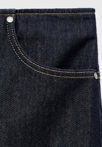 Donkere denim jeans met een voorzak omlijnd in contrasterende gele stiksels, met een metalen knoopaccent aan de bovenkant. Glad textuur.