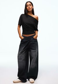 Fekete váll nélküli felső, összegyűrt textúrával, széles szárú, kifakult fekete farmerral és világos színű sportcipővel párosítva.