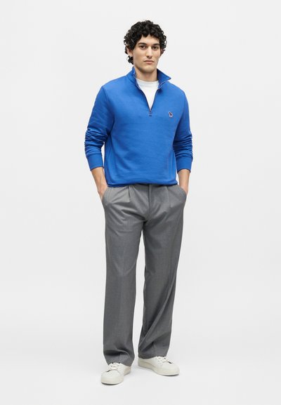 Νέος άνδρας στέκεται με τα χέρια στις τσέπες, φοράει μπλε πουλόβερ με φερμουάρ μέχρι το στήθος, γκρι παντελόνι, λευκά αθλητικά παπούτσια, σε μονόχρωμο λευκό φόντο.