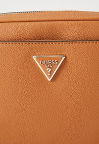 Oranssi teksturoitu tekonahkabag, jossa on hopeinen kolmion muotoinen emblem, jossa lukee "GUESS USA" ja keskellä kysymysmerkki.