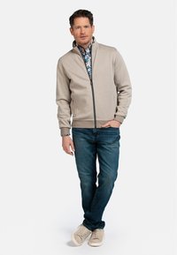 Beige zip-up jack met ribgebreide manchetten, gedragen over een gemarmerd blauw shirt, gecombineerd met blauwe jeans en lichtgekleurde sneakers.