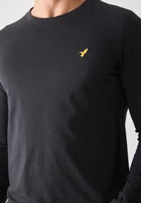 Must musta pikkade varrukatega T-särk, mis on valmistatud puuvillast; vasakul rinnaku piirkonnas on väike kollane tikandiga linnu logo; sile tekstuur.