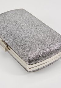 Pochette rectangulaire argentée pailletée avec un cadre métallique et une fermeture à pince sur un fond clair uni.