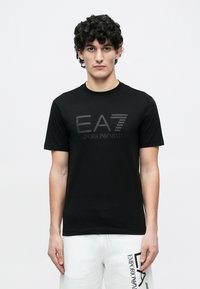 Göndör hajú férfi fekete EA7 Emporio Armani pólóban és fehér rövidnadrágban fekete logóval, egyszínű világos háttér előtt.