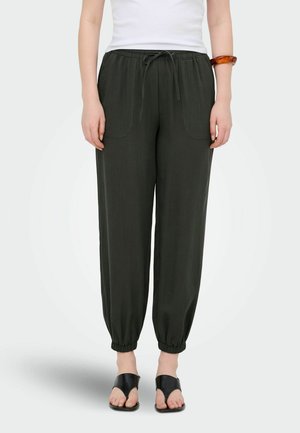 Vrouw draagt zwarte broek met trekkoord en elastische manchetten, wit mouwloos topje, zwarte sandalen en amber armband aan de rechterpols.