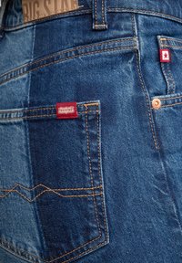 Denim jeans met een patchworkdesign in verschillende tinten blauw, oranje stiksels en rode merketiketten op de zakken.