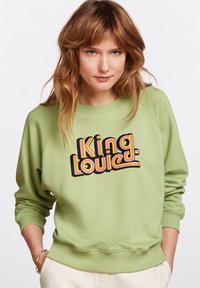 Felpa verde con scollatura tonda e maniche lunghe, caratterizzata da una grande grafica in stile retro che dice "King Louie" in colori vivaci.