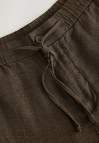 Gros plan d'un pantalon en tissu marron texturé avec une ceinture élastique, une fermeture par bouton et un lacet noué à l'avant.