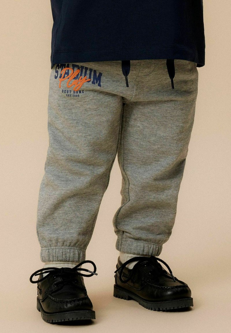 Grijze sweatpants met elastische boorden, voorzien van een blauwe en oranje "Stadium Play" print. Gecombineerd met zwarte veterschoenen. Zachte stof textuur.