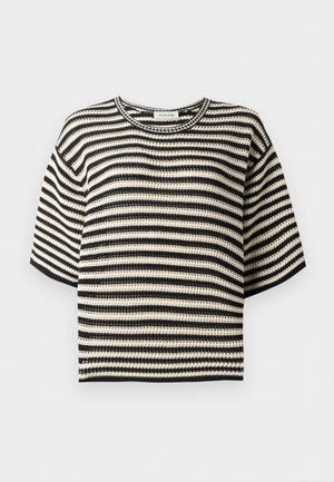 Kobieta ubrana w czarno-biały paskowany krótki top z krótkim rękawem, białe szerokie spodnie, czarne baleriny, trzyma dużą czarną skórzaną torbę typu tote.