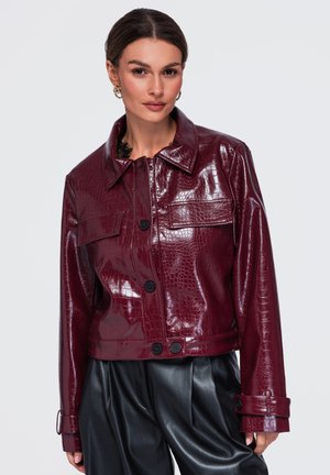 Femme portant une veste courte brillante en cuir synthétique crocodile rouge foncé avec des boutons noirs et un pantalon en cuir synthétique noir.