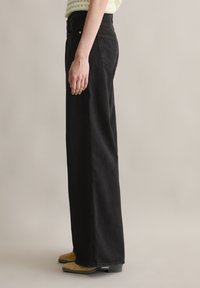Svarte vide jeans med høy midje, med synlige sømmer, frontlommer og en glatt tekstur. Komplimentert med beige sko.