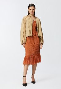 Bež bomber jakna do struka preko narančaste plisirane haljine s čipkastim rubom. Crne sandale na visoku petu upotpunjuju outfit.